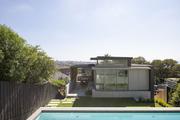 A contemporary two-storey residence viewed from the backyard, featuring a flat-roofed form, extensive floor-to-ceiling glazing, pale vertical cladding, and a covered outdoor circulation or entertaining area. A rectangular swimming pool occupies the foreground, with a compact lawn, stepping-stone path, timber privacy fencing, and mature trees surrounding the house. A distant urban hillside is visible beyond the property.