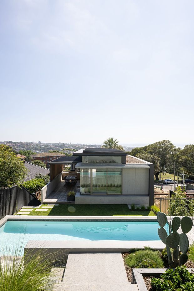 A contemporary multi-level residential house viewed from an elevated outdoor terrace, with a long swimming pool in the foreground, landscaped garden beds, concrete paving, lawn, and a modern flat-roofed glazed pavilion. The home uses dark cladding, concrete surfaces, extensive glazing, and layered roof forms, with a suburban hillside visible beyond.