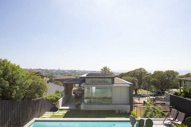 Exterior view of a contemporary multi-level residence on a sloping suburban site, photographed from an elevated rear terrace. The house features a flat roof, extensive glazing, pale rendered walls, dark boundary fencing, landscaped stepping stones, and a swimming pool in the foreground. A suburban urban landscape is visible beyond the property.