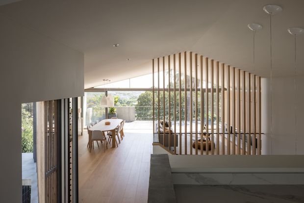 A bright contemporary open-plan interior with a kitchen in the foreground, a dining area beyond, and a large glazed opening leading to an outdoor terrace. Vertical timber slats form a sculptural screen and partial stair balustrade, while pale timber flooring, white walls, and natural daylight create a warm, airy atmosphere.