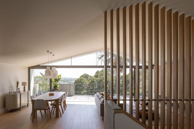 A bright contemporary open-plan dining and living area with warm timber flooring, white walls, and a full-width glazed opening to an outdoor terrace. A timber dining table and upholstered chairs sit beneath pendant lights, while a sofa is partially screened by a vertical timber batten partition. The space has leafy suburban views and abundant natural light.
