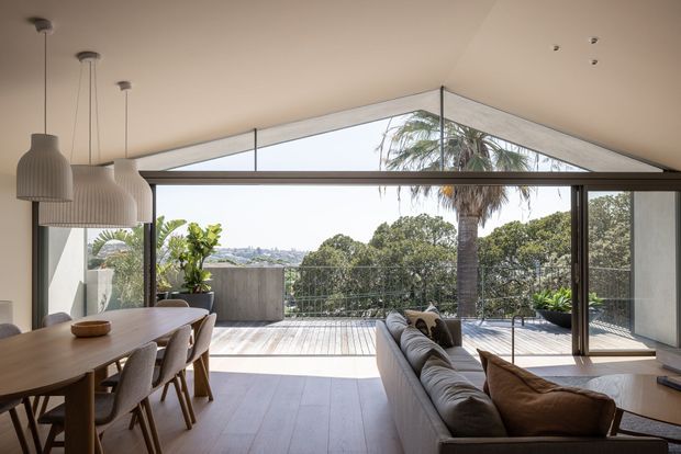 A bright, open-plan living and dining space with floor-to-ceiling glazing and a dramatic triangular gable window overlooking a leafy outdoor deck and palm trees. The room features a long timber dining table, upholstered dining chairs, a large neutral sofa, pendant lights, and pale timber flooring. The architecture has a refined contemporary feel with strong indoor-outdoor connection and abundant natural light.