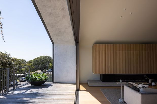 A refined indoor-outdoor living area with large sliding glass doors opening onto a timber deck and balcony railing. The interior features a warm neutral palette, a sculptural sectional sofa, a dark low media console, and a vertical timber-slatted wall feature framing the television. The space has strong natural light, clean contemporary lines, and views toward mature greenery and neighbouring homes.