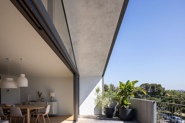 A bright contemporary apartment terrace or balcony viewed from an open indoor dining area. The space features a broad concrete soffit and side wall, full-height sliding glass doors with dark metal frames, a concrete balcony floor and balustrade, large potted plants, and an adjacent minimalist dining room with a timber table, upholstered chairs, and pendant lights. A leafy suburban or urban outlook is visible beyond the railing.