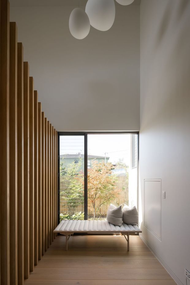 A refined, minimalist interior nook with a tall window overlooking a small garden courtyard. A built-in or fixed-looking upholstered bench sits beneath the glazing with two cushions, while a dramatic vertical timber batten screen lines the left wall and extends upward toward the ceiling. The space has pale timber flooring, smooth warm-white walls, slim dark window frames, and sculptural white pendant lights.