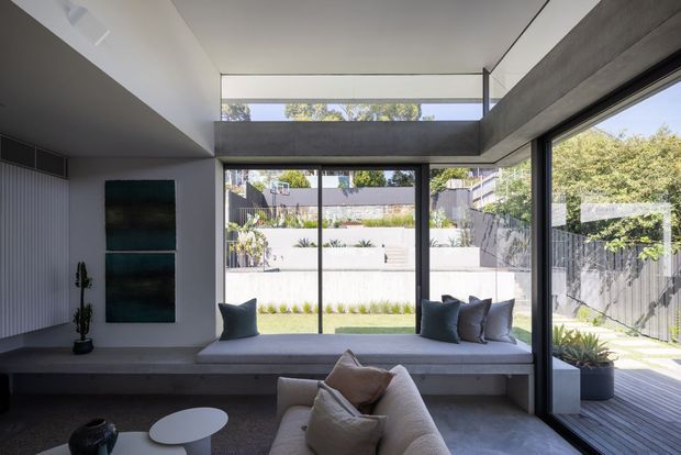 Contemporary living room with a built-in window seat, large sliding glass doors, a deep concrete or painted structural beam, and a high clerestory opening. The space opens directly onto a timber deck and landscaped courtyard, with neutral furnishings and strong indoor-outdoor connection.