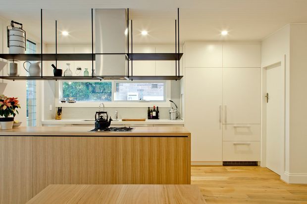 Bright contemporary kitchen with a large light-wood island and matching dining table, white cabinetry, timber flooring, a pale benchtop, and open black-framed shelving displaying glassware. A horizontal window provides natural light above the rear benchtop, with integrated appliances and a concealed pantry or storage wall on the right.