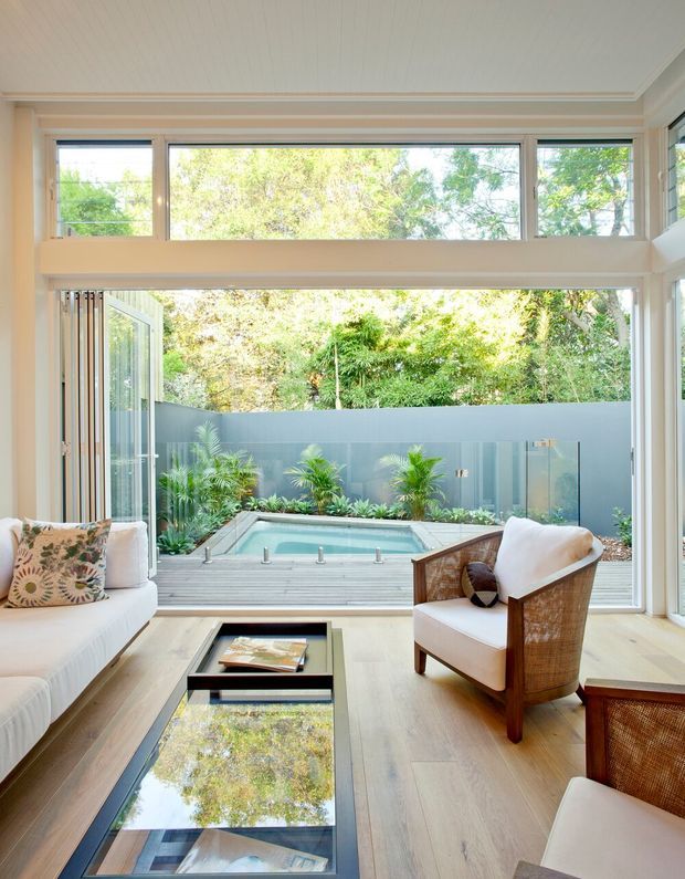 Bright contemporary living room opening onto a landscaped courtyard with a small plunge pool. The room features expansive floor-to-ceiling glazing, high clerestory windows, pale timber flooring, a light upholstered sofa, woven timber-framed armchairs, and a low black glass coffee table. The space has abundant natural light and a strong indoor-outdoor connection.