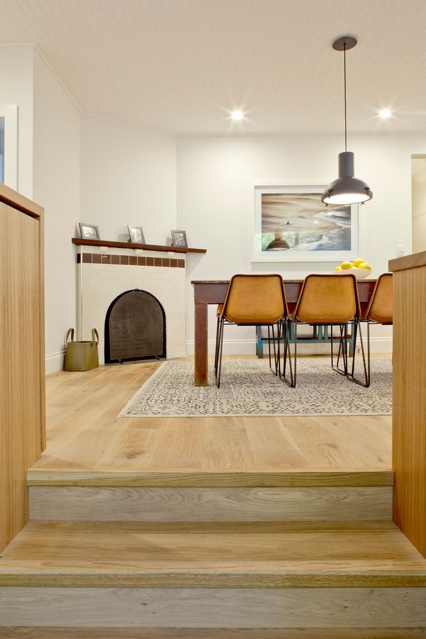 A warm, refined dining area viewed from a lower hallway through two broad timber steps. The room features pale timber flooring and stepped platforms, a white-painted fireplace with a dark arched fire screen and brown tile trim, a dark timber dining table, caramel leather dining chairs, a patterned neutral rug, framed coastal artwork, and a suspended contemporary pendant light. White walls and a white timber-lined ceiling create a bright, softly layered interior.