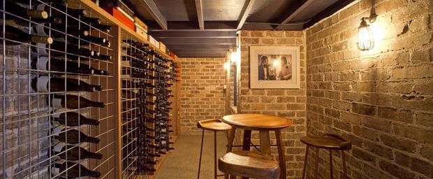 A compact, atmospheric wine cellar with extensive wall-mounted and freestanding bottle racks, exposed brick walls, dark timber ceiling beams, warm caged wall lighting, and a small round wooden table with stools. The space has a rustic, intimate and cellar-like character.