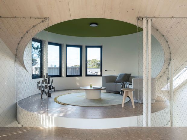 A bright, sculptural living area viewed through a curved opening and protective rope-netting. The room features a raised circular platform with a grey sectional sofa, round coffee table, pale rug, and small stool. Large black-framed windows provide natural light, while the curved timber-clad ceiling and green circular ceiling inset create a distinctive contemporary interior. A stair or split-level edge is visible in the foreground.