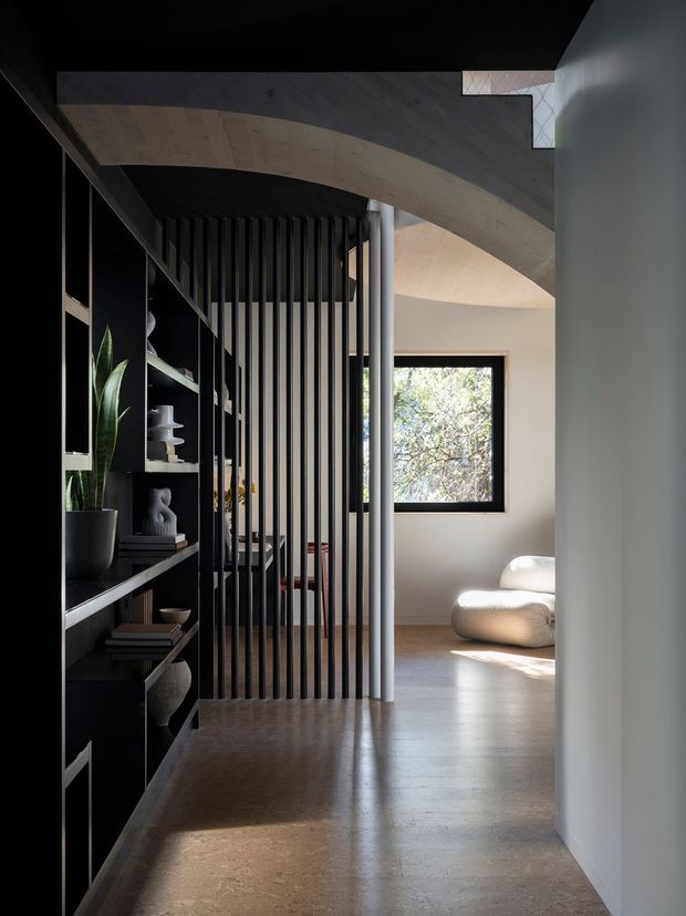 A moody contemporary interior hallway or transitional living area featuring a dramatic curved timber-lined ceiling, dark built-in shelving, and a black vertical-slat screen dividing the circulation space from a softly lit sitting area. Smooth polished concrete flooring, pale walls, sculptural cream seating, and a large window create contrast between the dark joinery and bright natural light. A tall indoor plant and curated objects are displayed on the shelving.