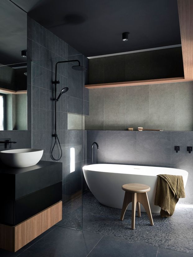A dark, contemporary bathroom with a freestanding white bathtub, wall-mounted black tapware, a separate open shower, vessel basin, and extensive charcoal-grey tiled surfaces. Black ceiling and dark cabinetry are balanced by warm timber drawer fronts and a timber shelf detail. A large mirror reflects the vanity and shower area, while a small stool and draped towel add limited furnishing.