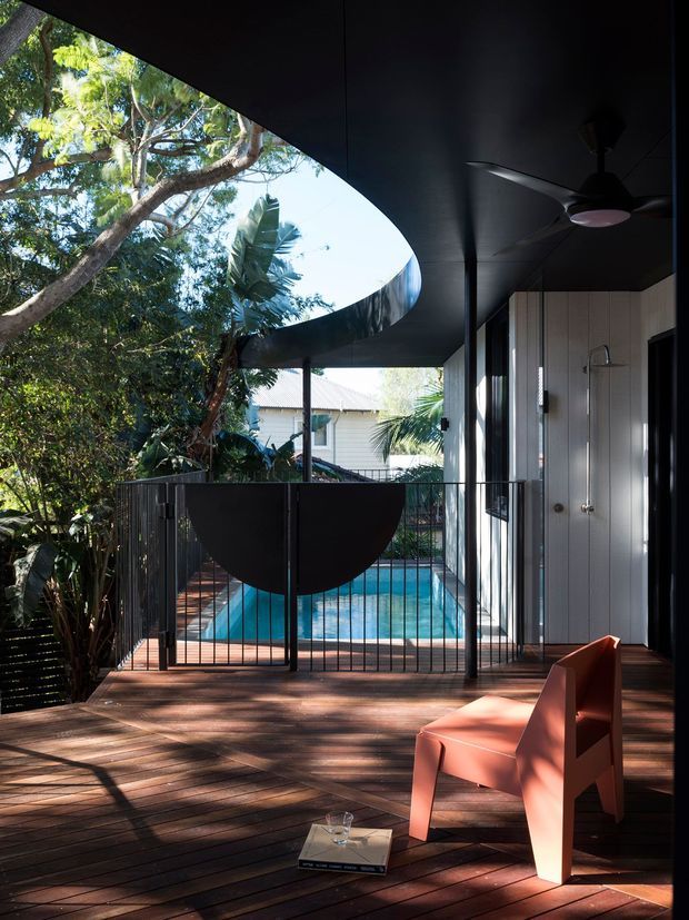 A tropical modern outdoor deck or covered terrace beside a swimming pool. The space features dark timber decking, a dramatic curved black ceiling and roof opening framing mature greenery, slender black metal balustrades, a white vertically clad wall with an exterior shower, ceiling fan, and a sculptural coral-red outdoor chair. The pool and lush vegetation are visible beyond the railing.