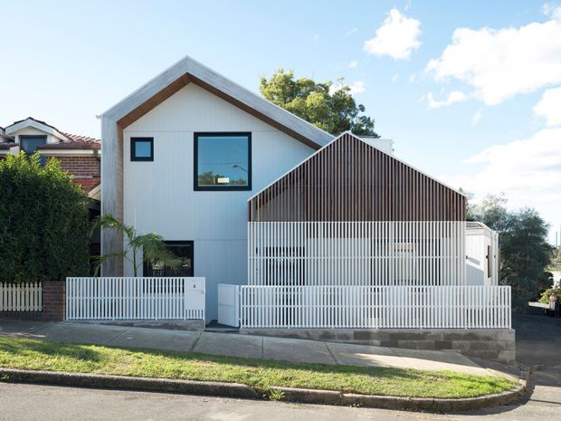 Street-facing view of a contemporary residential house with a gabled upper volume clad in light vertical panels and framed by warm timber trim. A projecting front structure features a steep gable, dark vertical timber battens, and a white vertical-bar screen behind a low masonry wall. White fencing, concrete paving, neighbouring homes, and established greenery complete the suburban streetscape.