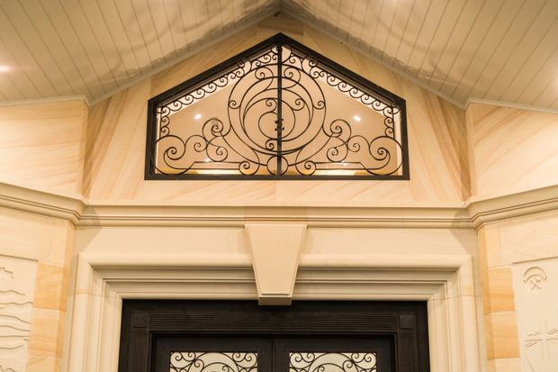 Close-up interior view of an ornate entryway or foyer, focused on a black decorative wrought-iron transom grille above a dark framed doorway. The space features warm cream and pale timber wall panelling, classical moulded trim, a steeply pitched timber-lined ceiling, and symmetrical carved detailing.
