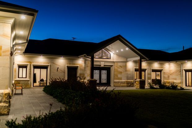 Night-time exterior view of a sprawling single-storey residence arranged around a landscaped courtyard. The home has pale stone or masonry-clad walls, dark pitched gable roofs, recessed downlighting, large glazed doors, decorative entry detailing, and a covered central porch supported by columns. Paved walkways and low planting surround a dark lawn, creating a formal, warmly illuminated facade.