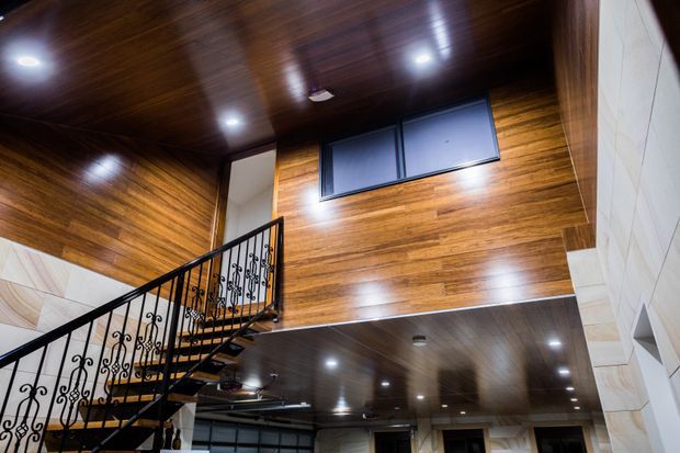 Interior architectural view looking upward toward a staircase and mezzanine/landing. The space features richly stained timber wall and ceiling panelling, recessed downlights, a dark-framed high window, and an ornate black wrought-iron balustrade. A lower level is visible beneath the landing, creating a strong vertical connection between floors.