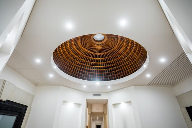 Interior architectural view looking upward at a bright white ceiling featuring a large circular domed timber feature with concentric radial slatted detailing and a central skylight or light fitting. Recessed downlights and linear ceiling panels surround the feature, with a tall corridor or entry space visible below.