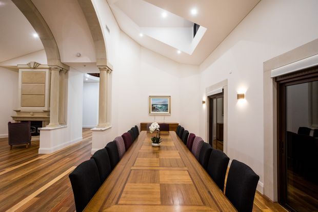 Formal dining room with a long rectangular timber dining table and upholstered dining chairs, set within a high-ceilinged interior. The room features polished parquet-style timber flooring, pale plaster walls, classical arched openings with decorative column details, dark timber-framed doors, wall sconces, recessed lighting, and a roof skylight or clerestory opening. A fireplace or decorative built-in feature is visible in the adjoining area.
