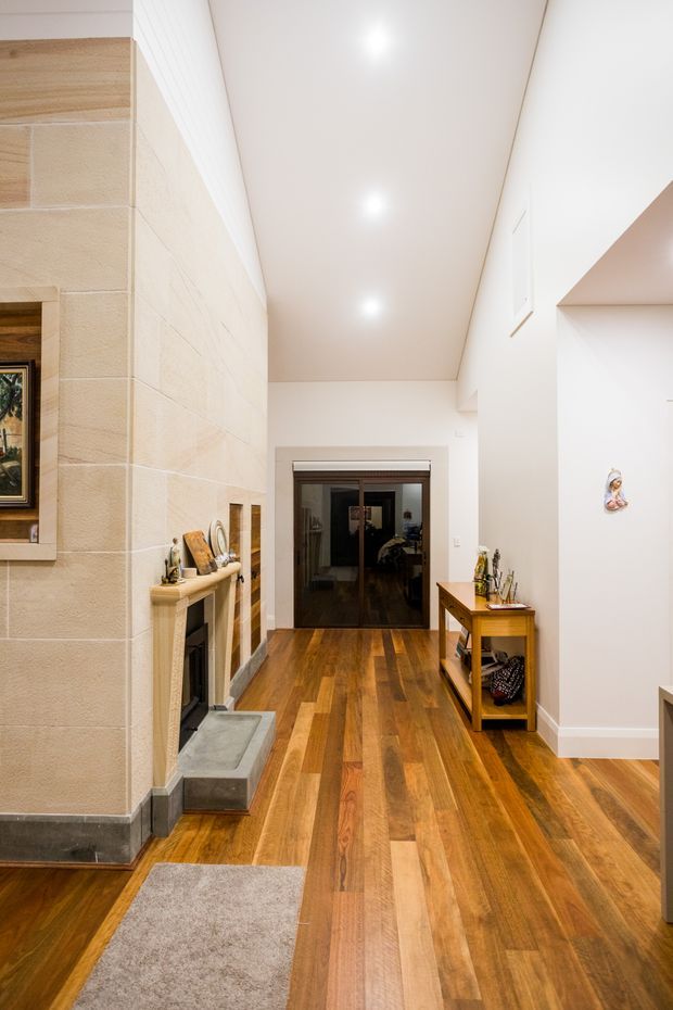A bright residential hallway or entry passage with warm timber flooring, white walls and ceiling, recessed downlights, and a large pale stone fireplace with a dark hearth on the left. A timber console table sits along the right wall, while glazed double doors lead to another room at the rear. The space has clean contemporary detailing with warm traditional materials and a high, elongated ceiling.