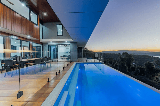 Contemporary multi-level home with extensive glazing, warm vertical timber cladding, and a dramatic cantilevered roof overlooking a tree-covered landscape. A long illuminated swimming pool extends along the elevated terrace, bordered by frameless glass balustrades and large-format paving. The image is captured at dusk, creating a refined blue-and-warm-light atmosphere.