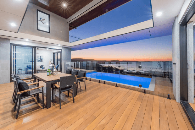 A contemporary indoor-outdoor entertaining area opening onto a timber deck with a vivid blue swimming pool and expansive waterfront or coastal views at sunset. The space includes a dining table with upholstered chairs, extensive floor-to-ceiling glazing, a timber-lined ceiling section, exposed structural beams, and large sliding glass doors.