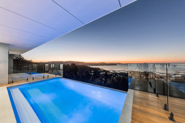 Modern rooftop or elevated outdoor swimming pool with vivid blue water, framed by glass balustrades and a broad projecting ceiling plane. The view overlooks a coastal or waterfront landscape at sunset, with a sleek contemporary architectural setting and timber-toned terrace decking.