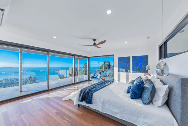 Bright contemporary bedroom with a large bed positioned beside expansive corner glazing, offering panoramic coastal and ocean views. The room has white walls and ceiling, warm timber flooring, blue and white bedding and cushions, recessed lighting, a timber-bladed ceiling fan, and a textured grey upholstered bedhead. Artwork and reflective decorative elements appear on the wall, while the glazing opens visually toward a balcony or terrace and the sea beyond.