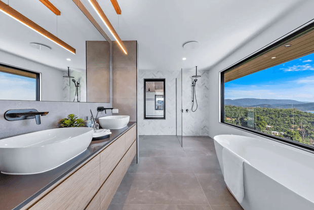 A spacious contemporary bathroom with a freestanding white bathtub positioned beside a large picture window overlooking a green, mountainous landscape. The room features a long double vanity with vessel basins, wall-mounted brushed-metal taps, light timber cabinetry, a dark benchtop, large grey floor tiles, marble-look wall surfaces, frameless glass shower screens, and linear timber-accent ceiling lighting.