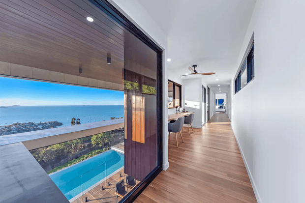 A bright contemporary residential hallway with warm timber flooring and a white wall, opening onto a balcony or terrace with expansive coastal views. The outdoor area includes a vivid blue swimming pool, while the interior includes a compact dining or breakfast area, timber ceiling detail near the glazing, recessed lighting, and a ceiling fan. Large dark-framed sliding glass doors connect the hallway to the outdoor space.