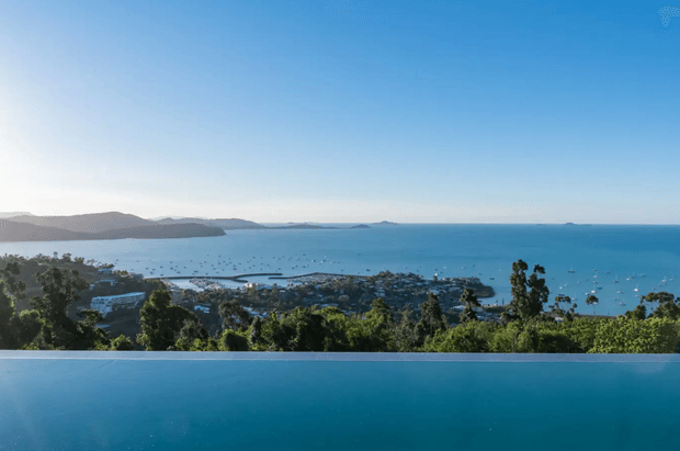 A wide panoramic coastal view from an elevated property, looking across a lush treed landscape and a marina-filled bay toward distant headlands. The lower portion of the image shows the edge and blue surface of an infinity pool merging visually with the sea. The scene is outdoors, bright, serene, and dominated by blue water and sky with dense green vegetation in the foreground.