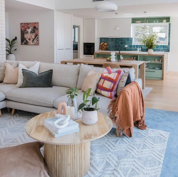Bright open-plan living room with a pale upholstered sofa, round timber coffee table, layered patterned rug, indoor plants, and colourful cushions. A compact contemporary kitchen is visible behind it, featuring white cabinetry, pale timber surfaces, open shelving, and a distinctive teal-blue tiled splashback. The space has abundant natural light and a relaxed, colourful coastal feel.