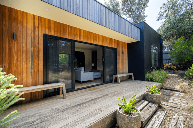 Exterior view of a contemporary single-storey home with a warm vertical timber-clad facade, dark metal upper cladding, and large black-framed sliding glass doors opening onto a broad timber deck. Simple timber benches, concrete planters, gravel landscaping, and established trees surround the entrance and outdoor living area.