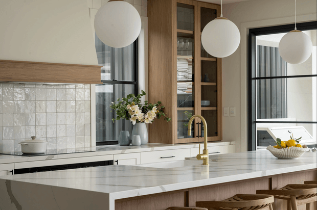 A bright, contemporary kitchen centered on a large marble-look island with an integrated sink and brushed brass gooseneck tap. The kitchen features white lower cabinetry, warm timber cabinetry and shelving, a textured square-tile splashback, black-framed glazing, and three large spherical pendant lights. Timber bar stools line the island, while a bowl of lemons and floral arrangement add restrained decorative accents.