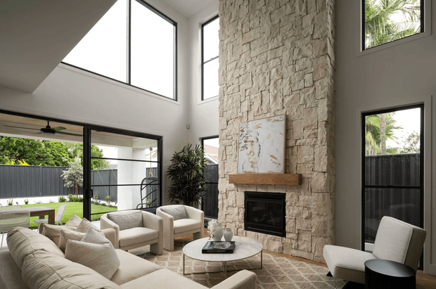 Bright double-height contemporary living room with expansive floor-to-ceiling glazing, a substantial textured stone fireplace wall, light upholstered seating, a round coffee table, and a large neutral patterned rug. The room opens visually to a landscaped outdoor area and includes an abstract artwork above the fireplace.