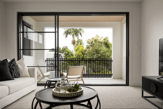 A bright, contemporary living room viewed toward a large open glazed doorway leading to a balcony or terrace. The room contains a pale sofa with black-and-white cushions, a round black coffee table, a low black media console, and a light woven lounge chair. The broad black-framed glazing opens to an outdoor balcony with a dark vertical railing and leafy trees beyond. A timber-lined soffit is visible outside, while the interior has light neutral walls and carpet.