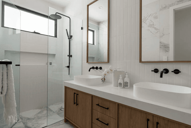 Contemporary bathroom featuring a long double vanity with two white vessel basins, warm timber cabinetry, a white benchtop, black wall-mounted tapware, framed mirrors, white vertical wall tiles, and a glass-enclosed walk-in shower. Marble-look floor and shower tiles add subtle grey veining, while high windows provide natural light.