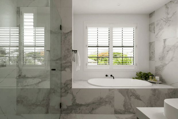 Bright, refined contemporary bathroom featuring a freestanding oval bathtub set into a marble-clad surround beneath a shuttered window. A frameless glass shower enclosure occupies the left side, with matching marble-look wall and floor surfaces throughout. A partial white vanity and basin are visible at the lower right, while greenery and outdoor planting are seen through the window.
