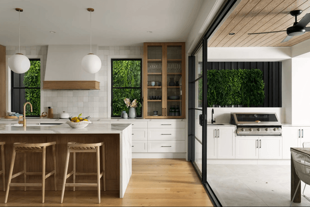 A bright, open-plan kitchen with a large white island, timber bar stools, white tiled splashback, timber rangehood shelf, brass tapware, and built-in white cabinetry. Glazed steel-framed doors separate the kitchen from a covered outdoor barbecue area with matching cabinetry and a ceiling fan. The kitchen includes a timber display cabinet, pale timber flooring, pendant globe lights, and views to lush greenery.