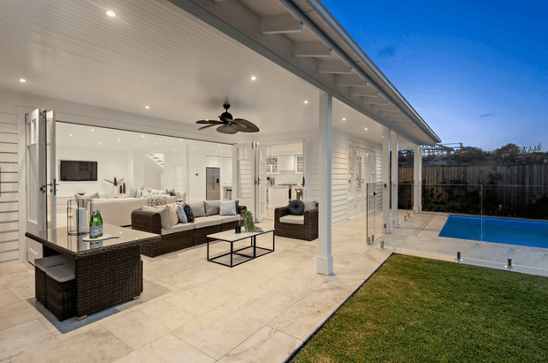 A bright, covered outdoor entertaining area opens onto a lawn and swimming pool. The space features white horizontal wall cladding, a white-lined ceiling with recessed downlights, ceiling fans, large glazed folding doors, pale tiled flooring, and a wicker outdoor lounge and bar setting. The interior living area is visible through the open glazing, creating a seamless indoor-outdoor connection.