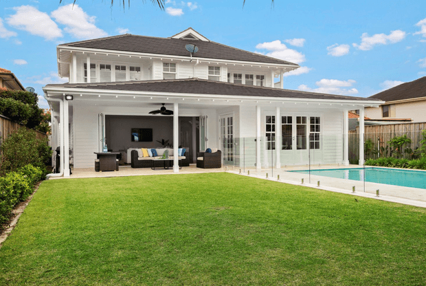 A bright rear exterior view of a two-storey white weatherboard-style home with a broad covered outdoor living area, louvred upper-level windows, French doors, and a glass pool fence. A turquoise swimming pool sits beside the patio, opening onto a large manicured lawn under a blue sky.