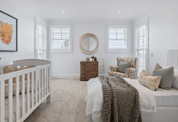 Bright, calm nursery-bedroom with white plantation-shuttered windows, pale carpet, a white cot, upholstered bed, vintage chest of drawers, wingback armchair, round wall mirror, and soft neutral textiles. The room has a refined coastal-traditional feel with abundant natural light and restrained beige, grey, and white tones.