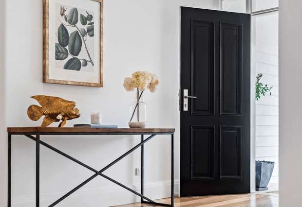 A refined residential entryway with a black paneled interior door, white walls and trim, and warm timber flooring. A slim dark wood console table with black metal cross-braced legs holds decorative objects, while botanical artwork and a vase of dried flowers add natural accents. A bright exterior or adjoining space is visible through the open doorway.