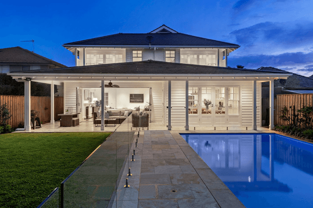 A large two-storey suburban residence viewed from the backyard at dusk. The home has a broad low-pitched tiled hipped roof, white rendered or weatherboard-style walls, extensive glazed doors and windows, and a covered outdoor entertaining veranda. A paved path leads through a manicured lawn toward the house, with a rectangular blue swimming pool along the right side. The warmly illuminated interior is visible through the glazing, including a living area and built-in cabinetry.
