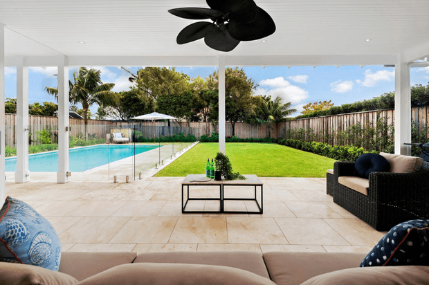 Covered outdoor entertaining area opening onto a backyard with a swimming pool and lawn. The space has a tiled patio, white structural posts, extensive frameless glass pool fencing, a ceiling fan, outdoor wicker seating, and a low metal coffee table. Tropical planting and timber fencing are visible beyond the yard.