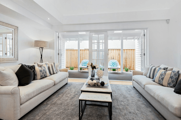 Bright, airy living room with two large neutral sofas facing each other across nested coffee tables and a textured grey rug. White walls and ceiling, generous glazed doors or windows opening to a timber-fenced outdoor area, patterned blue-and-white cushions, and a large framed mirror create a relaxed coastal feel.