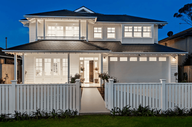 Two-storey suburban residence photographed at dusk, with a symmetrical white-painted facade, dark tiled hipped roof, broad eaves, shuttered windows, covered upper balcony, ground-floor entry porch, and an integrated double garage. A white picket-style fence and landscaped planting frame the front elevation.
