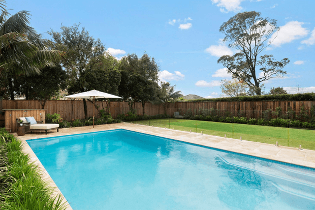 A bright outdoor swimming pool occupies the foreground, surrounded by pale paved coping, manicured lawn, dense planting, and a timber privacy fence. A white poolside umbrella and lounge seating sit to the left, with mature trees and a clear blue sky beyond. The scene has a relaxed suburban resort-like character.