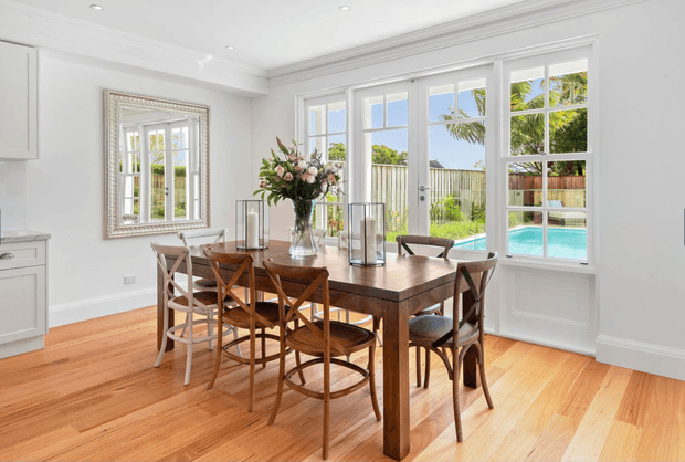 Bright formal dining room with a large dark timber dining table and mixed wood dining chairs, positioned beside expansive white-framed French doors and windows overlooking a swimming pool and garden. The room features white walls, decorative ceiling cornices and skirting, warm timber flooring, a mirrored wall, and a floral centrepiece with glass candle lanterns.